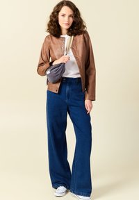Giacca di pelle marrone con chiusura a scatto, camicia bianca, pantaloni di jeans blu larghi e borsa a tracolla grigia. Modello con sneakers bianche.