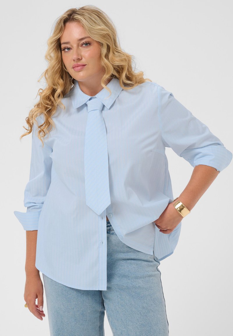 Femme blonde portant une chemise bleu clair avec une cravate assortie, manches retroussées, jean clair et un bracelet en or, posant avec une main sur la hanche.