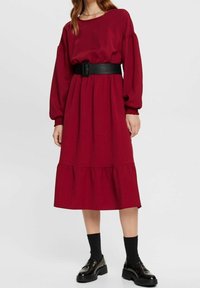 Robe rouge à manches longues avec un ourlet en couches, tissu texturé, taille cintrée avec une large ceinture noire, accompagnée de chaussettes noires et de chaussures épaisses.