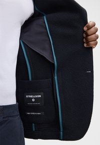 Dunkelmarineblaues Jacket aus strukturiertem Stoff, mit kontrastierender Türkis-Bordüre und Innentasche. Die Etiketten geben die Marke "Strellson" und Materialdetails an.