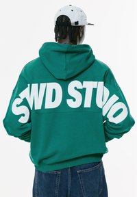 Person med fletninger iført en grøn hoodie med stor hvid "SWD STUVO" tekst på ryggen og en hvid baseballcap vendt væk.
