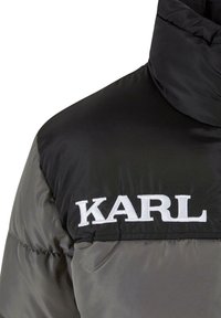 Puffjakke med en sort overdel og en grå underdel, med en høj krave og broderet logo "KARL" i hvid.