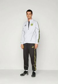 Balts sporta mētelis ar melniem akcentiem un zaļu logo, kombinēts ar tumšiem biksēm ar dzeltenām sānu svītrām. Uzvilktas melnas kurpes.