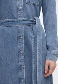 Κοντινή λήψη ενός φόρεματος από τζιν με μεταλλικά κουμπιά μπροστά, μακριά μανίκια, ζώνη από το ίδιο ύφασμα και εμφανείς ραφές.