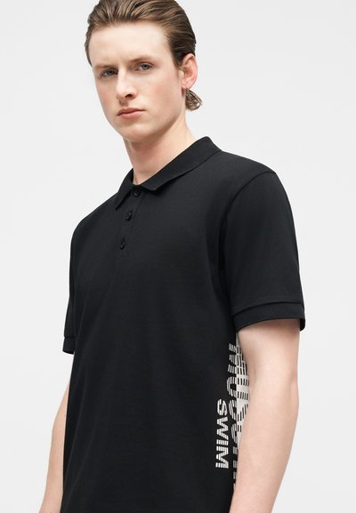 Jeune personne aux cheveux plaqués en arrière, portant un polo noir avec un texte blanc en diagonale sur le côté droit.