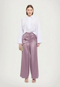 Egy nő áll, fehér hosszú ujjú blúzt és magas derekú, bő szárú, levendula színű selyem nadrágot visel virágmintával, arany clutch táskát tart a kezében.