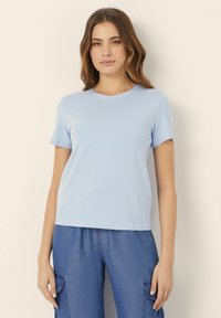 Tricou scurt cu mânecă scurtă de culoare albastru deschis, cu guler rotund, fabricat dintr-un material moale, asociat cu pantaloni de denim albaștri, lejeri, care au buzunare laterale.