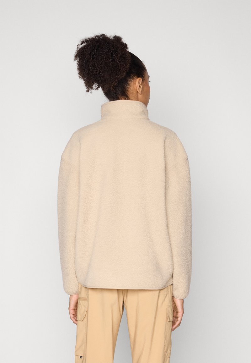 Frau mit lockigem Haar in einem hohen Dutt, trägt eine beige Fleecejacke und beige Cargohosen, von hinten vor einfarbigem Hintergrund gezeigt.