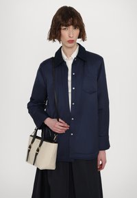 Mørkeblå button-up jakke med krave, båret over en hvid skjorte. Lyse beige håndtaske med sorte detaljer og dobbelte håndtag.