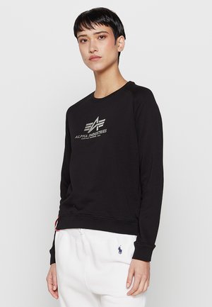Person mit kurzen dunklen Haaren, die einen schwarzen Alpha Industries Pullover und weiße Hose mit kleinem dunklem Logo auf der Tasche trägt.