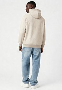 Sweat à capuche beige avec une capuche et des poignets côtelés, associé à un jean bleu clair à coupe ample. Texture lisse, détails minimalistes et baskets blanches.