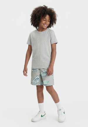 Niño sonriente con cabello rizado que lleva una camiseta gris lisa, pantalones cortos grises de Nike con logos verdes, calcetines blancos y zapatillas blancas con detalles verdes.