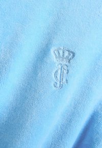 Material texturat albastru deschis, cu o coroană brodată și logo-ul literei „C” realizate dintr-un material moale și pufos.