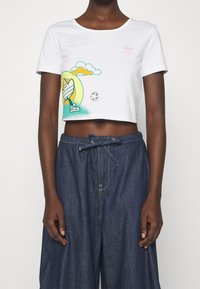 Vit kortärmad t-shirt med en färgglad surfbräda, gröna moln och en rosa logga; ihop med lösa mörkblåjeans-shorts.