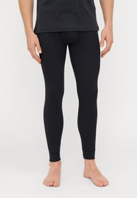 Μαύρες κολάν φτιαγμένες από ριγωτό ύφασμα, εφαρμοσμένος σχεδιασμός, σφηνοειδείς κορυφές και μανσέτες στα αστραγάλους. Φοριούνται με ένα μαύρο πουκάμισο, σε ανοιχτό φόντο.