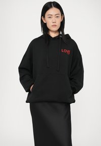 Crna pamučna hoodie majica s slobodnim krojem, koja ima crveni grafički natpis "LOVE" na prsima, kapuljaču s vezicom i kengursku džep. Glatka tekstura.