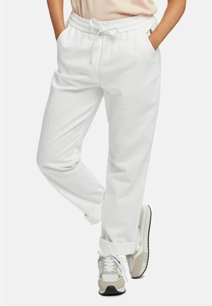 Personne portant un pantalon blanc à cordon avec les mains dans les poches et des baskets blanches et grises décontractées, debout avec une jambe croisée.