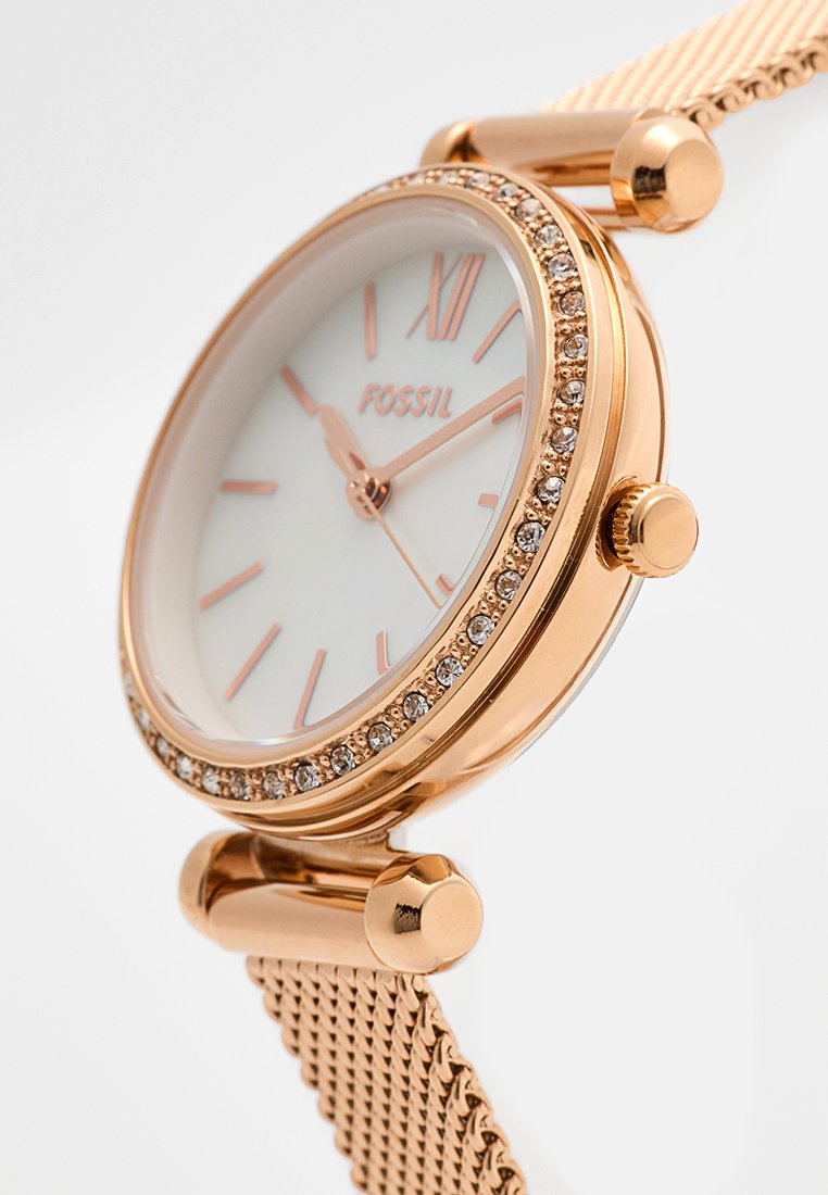 Roségoldene Uhr mit rundem Zifferblatt, weißem Ziffernblatt, roségoldenen Markierungen und Kristallelementen. Mesh-Armband mit glatter Oberfläche. Verfügt über eine Krone.