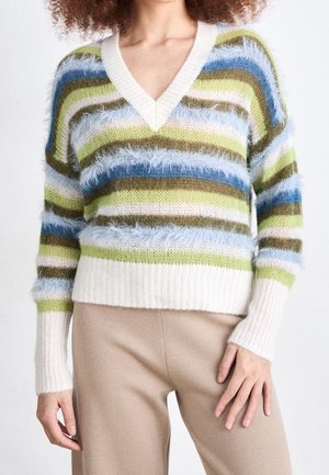 Femme portant un pull à col en V rayé et duveteux bleu, vert, marron et blanc avec un pantalon beige, présentée du cou aux hanches.