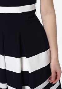 Ruka ženy spočívající vedle sukně černobíle pruhované plisované šaty, zobrazené od pasu po střed stehna na bílém pozadí.