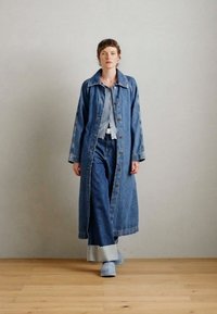 Cappotto di denim con design a colletto, chiusura a bottoni, maniche lunghe, abbinato a una camicia di un blu più chiaro e jeans a campana, tutti in diverse sfumature di denim.