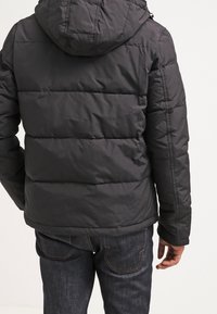 Czarna pikowana kurtka z kapturem, z poziomym wzorem pikowania, gładką teksturą i elastycznymi mankietami, połączona z ciemnymi dżinsami.