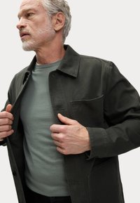 Olive green jacket with patch pockets, button closure, and collar, worn over a light green long-sleeve shirt. Textured fabric, casual design.