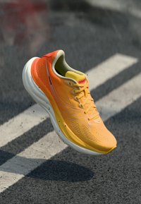 Φωτεινό πορτοκαλί και κίτρινο αθλητικό παπούτσι με λευκή σόλα, αιωρείται πάνω από άσφαλτο με λευκές οδικές σημάνσεις.