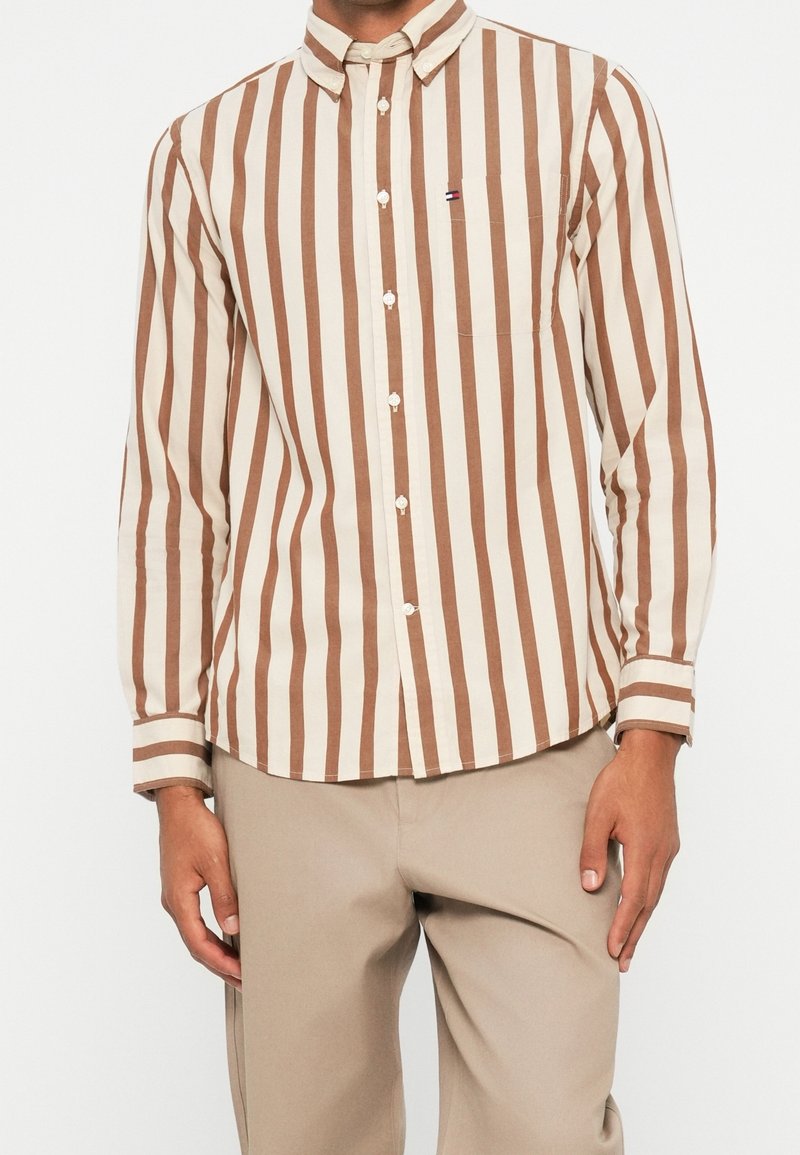 Homme portant une chemise à manches longues boutonnée avec des rayures verticales marron et blanches et un pantalon beige.