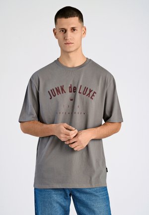 Camiseta de algodón gris con un estampado burdeos que dice "JUNK de LUXE" junto a un gráfico de una corona. Ajuste relajado y mangas cortas.