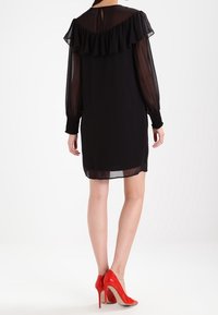 Femme portant une robe noire à manches transparentes avec un détail de volants dans le dos et des chaussures à talons hauts rouges vives, debout de dos.