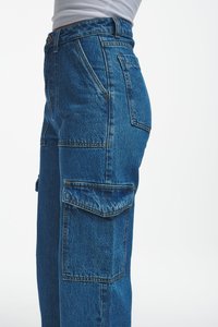 Blue high-waist denim cargo pants with large flap pockets on the thigh and side, worn with a tucked white top, side view.