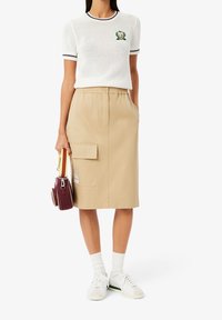 Vit kortärmad tröja med marinblå randig krage och broderad logotyp, tillsammans med en beige knälång kjol med sidofickor.