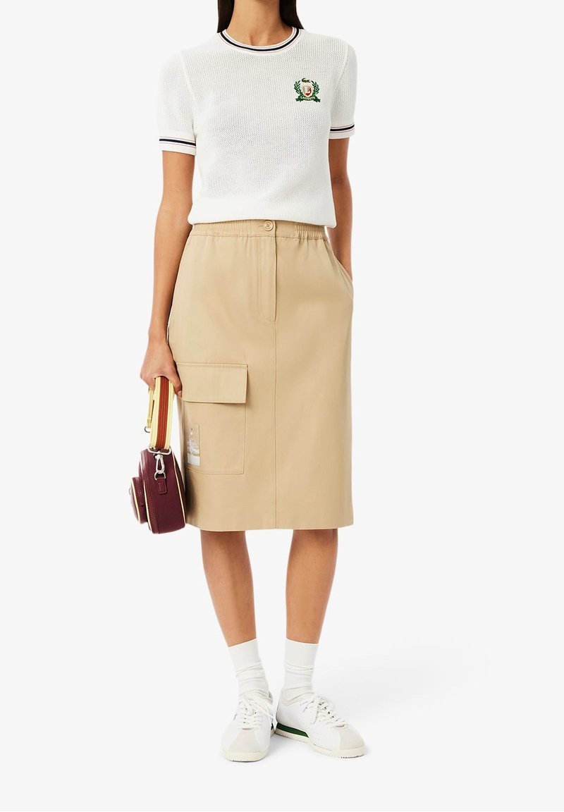 Vit kortärmad tröja med marinblå randig krage och broderad logotyp, tillsammans med en beige knälång kjol med sidofickor.