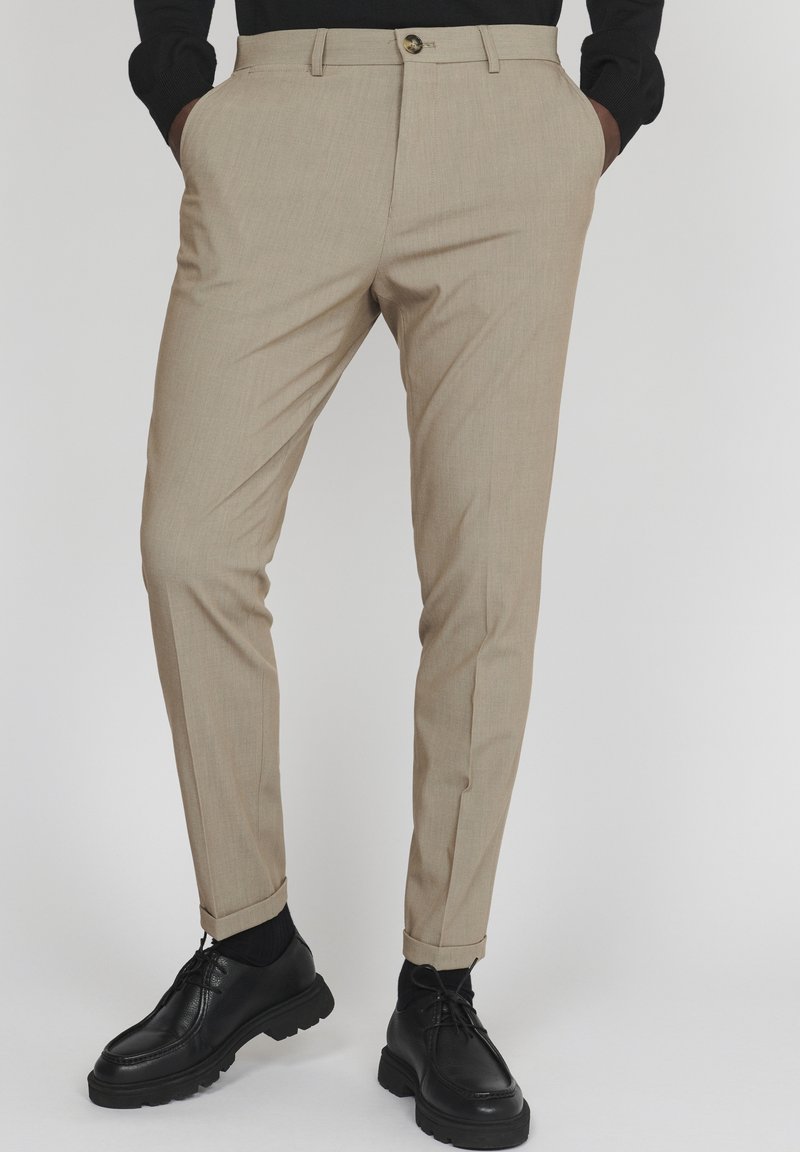Pantalon beige sur mesure avec ourlets retroussés, porté avec des chaussettes noires, des chaussures noires à lacets et un haut noir à manches longues avec les mains dans les poches.