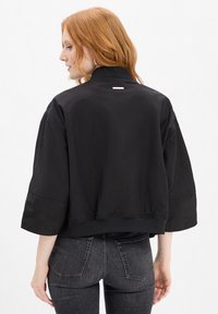 Czarna krótka kurtka z wysokim kołnierzem, szerokimi rękawami i elastycznym dołem. Gładki materiał z małym logo z tyłu.