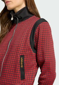 Röd houndstooth-zipjacka med svart ribbad krage och axelaccenter, med en orange logotagg på framlappen.