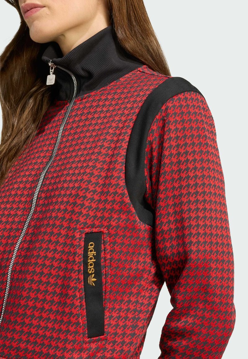 Giacca zip-up rossa a motivo houndstooth con colletto a coste nero e accenti sulle spalle, caratterizzata da un'etichetta arancione sulla tasca frontale.