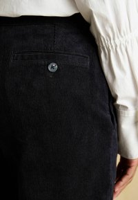 Navy zila ribota auduma bikses ar faktūru, aizmugurējo kabatu ar vienu melnu pogu un pieguļošu jostasvietu ar ruļļotu detaļu.