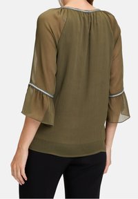 Blouse vert olive avec manches cloche en voile transparent et texture subtile. Présente un bord argenté autour du col. Portée avec un pantalon noir.