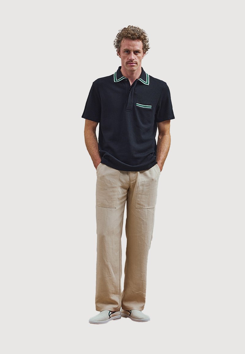 Zwarte polo met contrasterende groene kraag en zak, gecombineerd met lichtbeige broeken en grijze instappers. Casual ontwerp, ontspannen pasvorm.