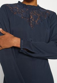 Mörkblå klänning med en blommig spetspanel vid halsen, volangkrage och långa  ärmar med veckade manschetter, tillverkad av mjukt tyg.