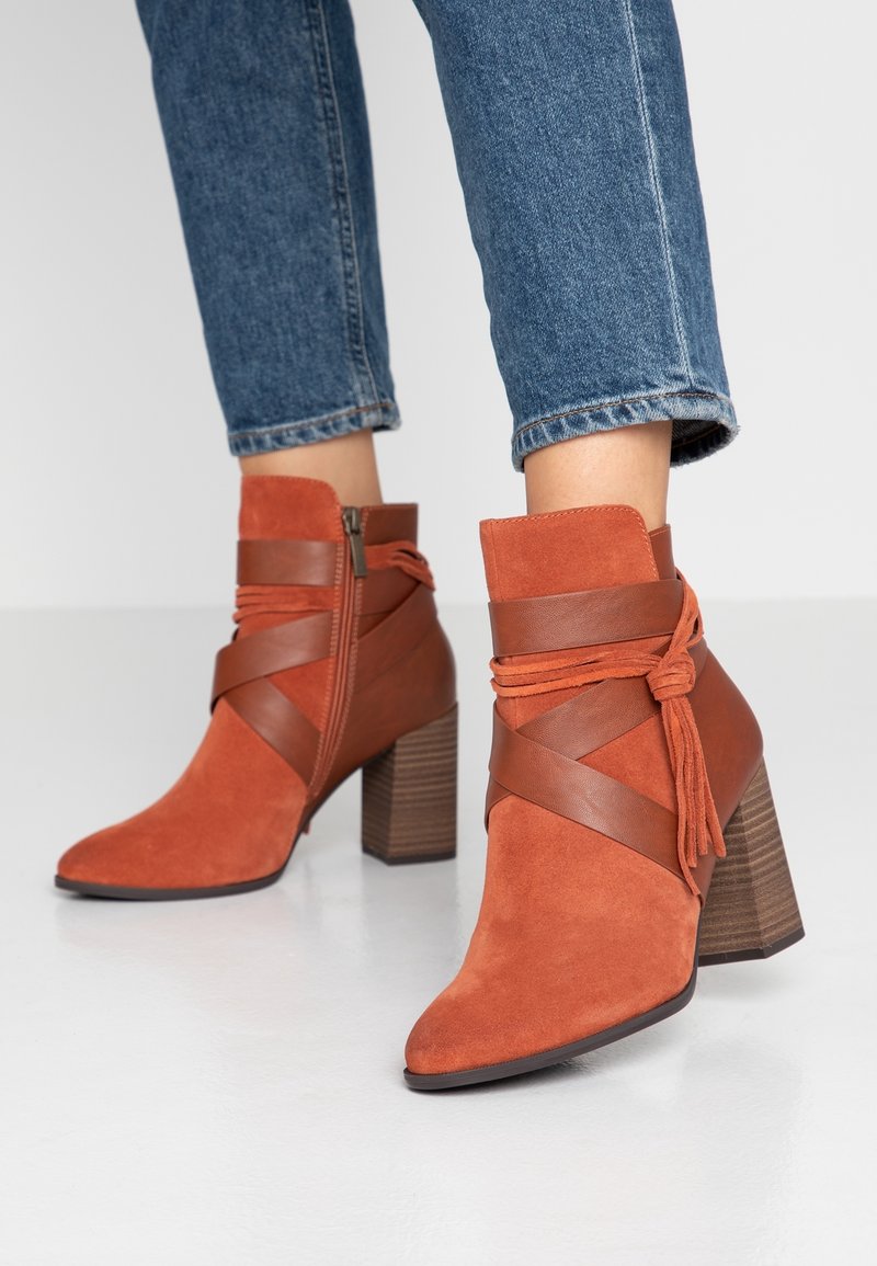 Bottes basses en daim couleur rouille avec des lanières en cuir marron et des talons carrés, portées avec un jean bleu sur un fond uni.