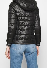 Sort pufferjakke med hætte, quiltet design, blank overflade og ribbede manchetter med Calvin Klein-logoet, parret med lyseblå jeans.