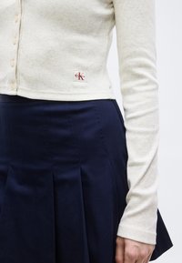 Μπεζ ριμπ πλεκτή μακρυμάνικη μπλούζα με κουμπιά και λογότυπο Calvin Klein, σε συνδυασμό με μπλε σκούρα πλισέ φούστα, που δείχνει τη μέση και το χέρι.