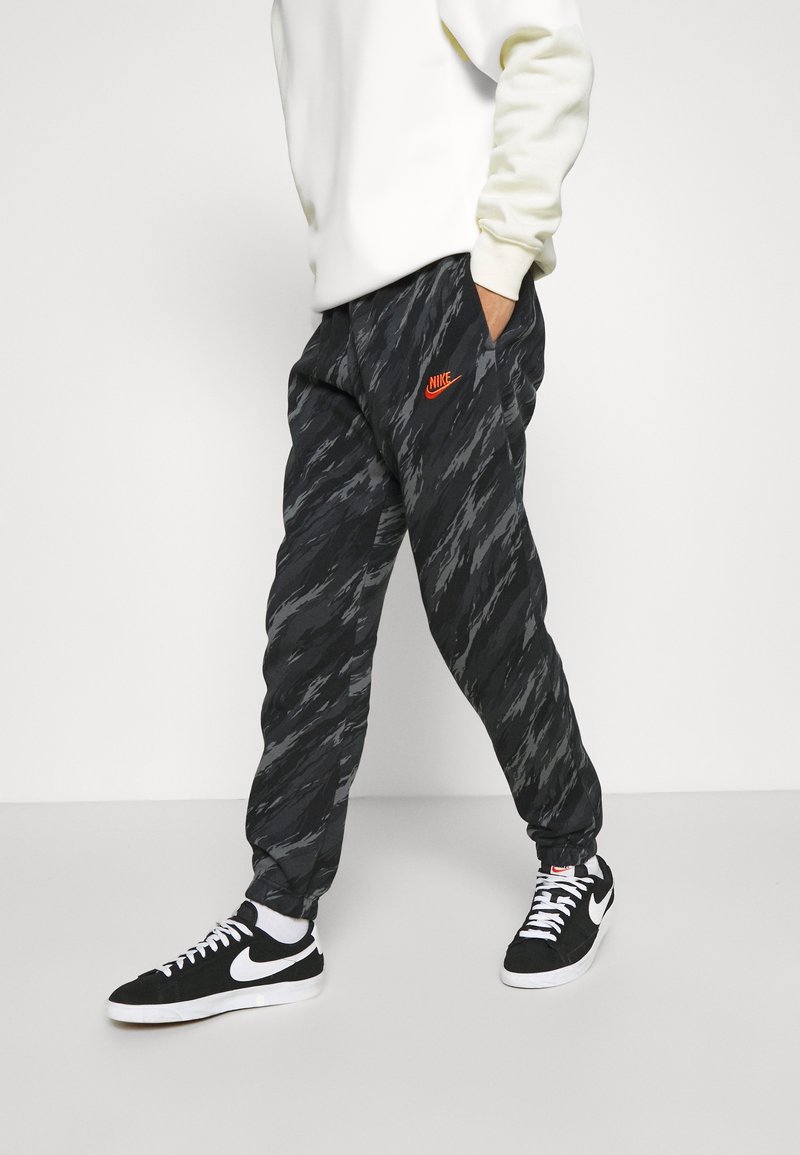 Svarta joggers med vågmönster och orange Nike-logotyp. Bärs med svarta sneakers och har ribbade muddar samt sidofickor.