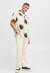 Camicia beige a maniche corte con motivo a sole nero, abbinata a pantaloni color crema e sneakers nere con dettagli bianchi.