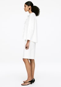 White, ruffled blouse with long sleeves, paired with a knee-length white skirt. Black, flat sandals complete the outfit.