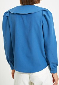 Blusa blu a maniche lunghe con un ampio colletto, spalle arricciate e polsini con bottoni. Tessuto liscio con una leggera lucentezza.