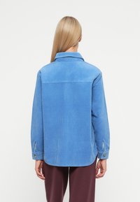 Ανοιχτό μπλε πουκάμισο κορουρόι με γιακά, μακριά μανίκια και κουμπωμένα μανίκια; η πίσω όψη δείχνει υφή κατακόρυφης ραβδωτής και κυματιστό στρίφωμα.